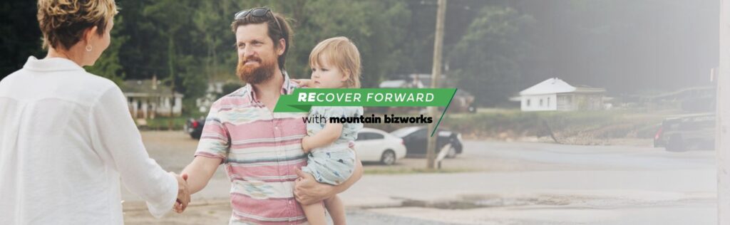 A Mountain BizWorks representative shakes hands with a small business owner holding a child, symbolizing community support and long-term small business recovery.
