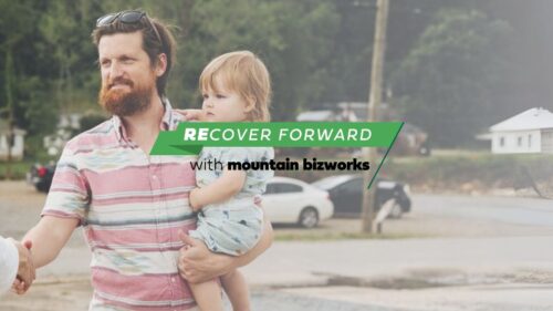 A Mountain BizWorks representative shakes hands with a small business owner holding a child, symbolizing community support and long-term small business recovery.
