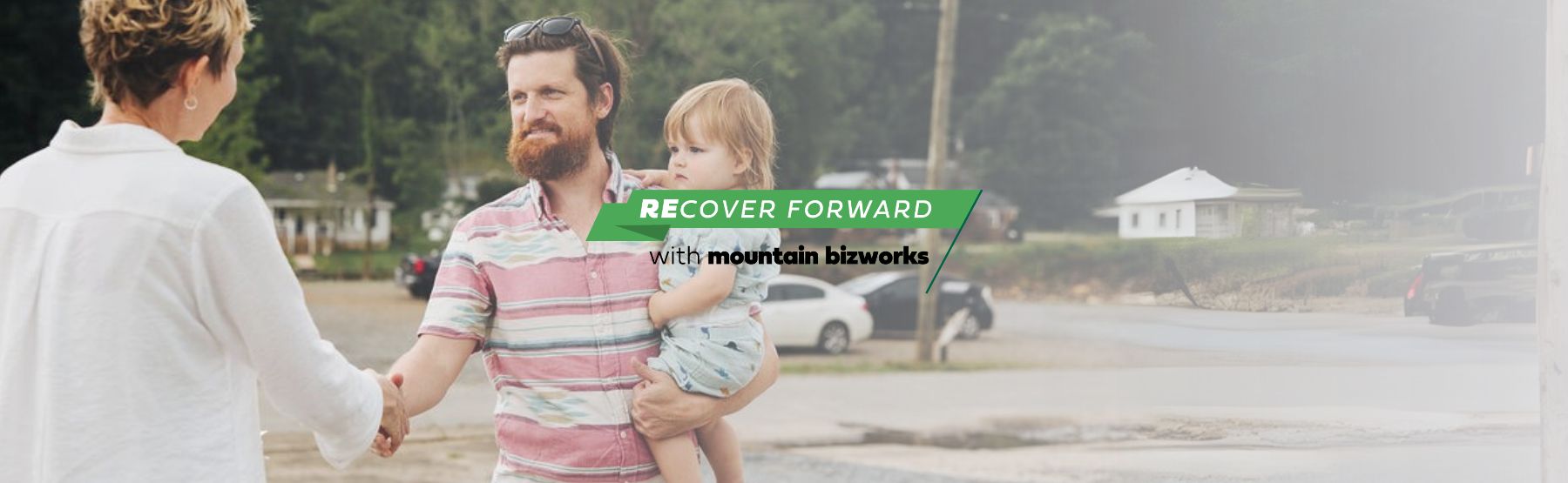A Mountain BizWorks representative shakes hands with a small business owner holding a child, symbolizing community support and long-term small business recovery.