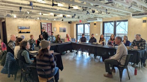 A diverse group of approximately 15-20 people at Mountain BizWorks' Waypoint Resilience Cohort Kick-off sit in a U-shaped arrangement of tables covered with black tablecloths in an industrial-style meeting space with exposed ceiling, concrete floors, and large windows. A facilitator in a tan shirt gestures while speaking to the engaged attendees, who have laptops, notebooks, and coffee cups in front of them.