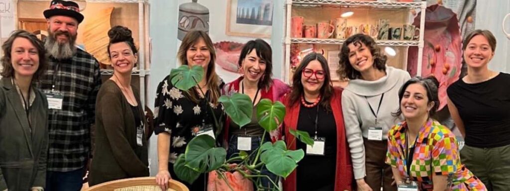The Craft Your Commerce cohort poses together in their booth at American Craft Made Baltimore, smiling with shelves of handmade ceramics and artwork displayed behind them.