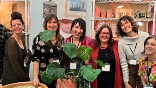 The Craft Your Commerce cohort poses together in their booth at American Craft Made Baltimore, smiling with shelves of handmade ceramics and artwork displayed behind them.