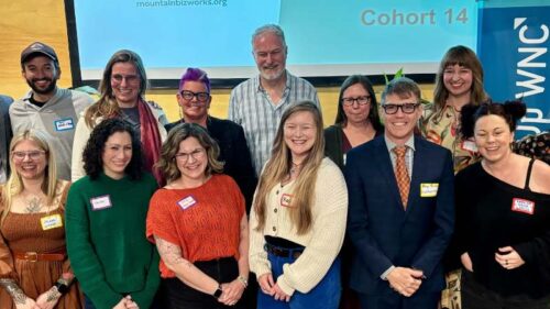 Group photo of the 14th ScaleUp WNC Cohort, approximately 17 participants posing together in two rows in front of a projection screen displaying 'mountainbizworks.org' and 'Cohort 14.' A blue ScaleUp WNC banner is visible on the right side. Many participants are wearing name tags. The group is diverse in age and appearance, smiling at the camera in what appears to be an indoor event or classroom setting.